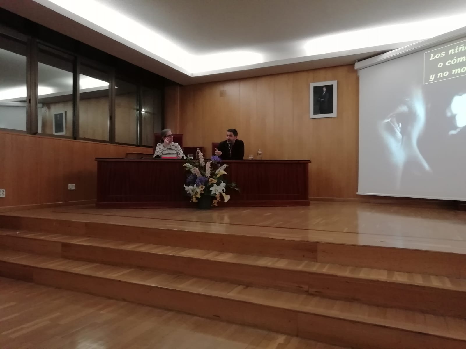 Imagen en la que se ve al profesor Serafín Béjar y a la ponente Ianire Angulo sentados durante la conferencia "Si no os hacéis como niños", a la derecha de ellos se puede observar una pantalla de proyección