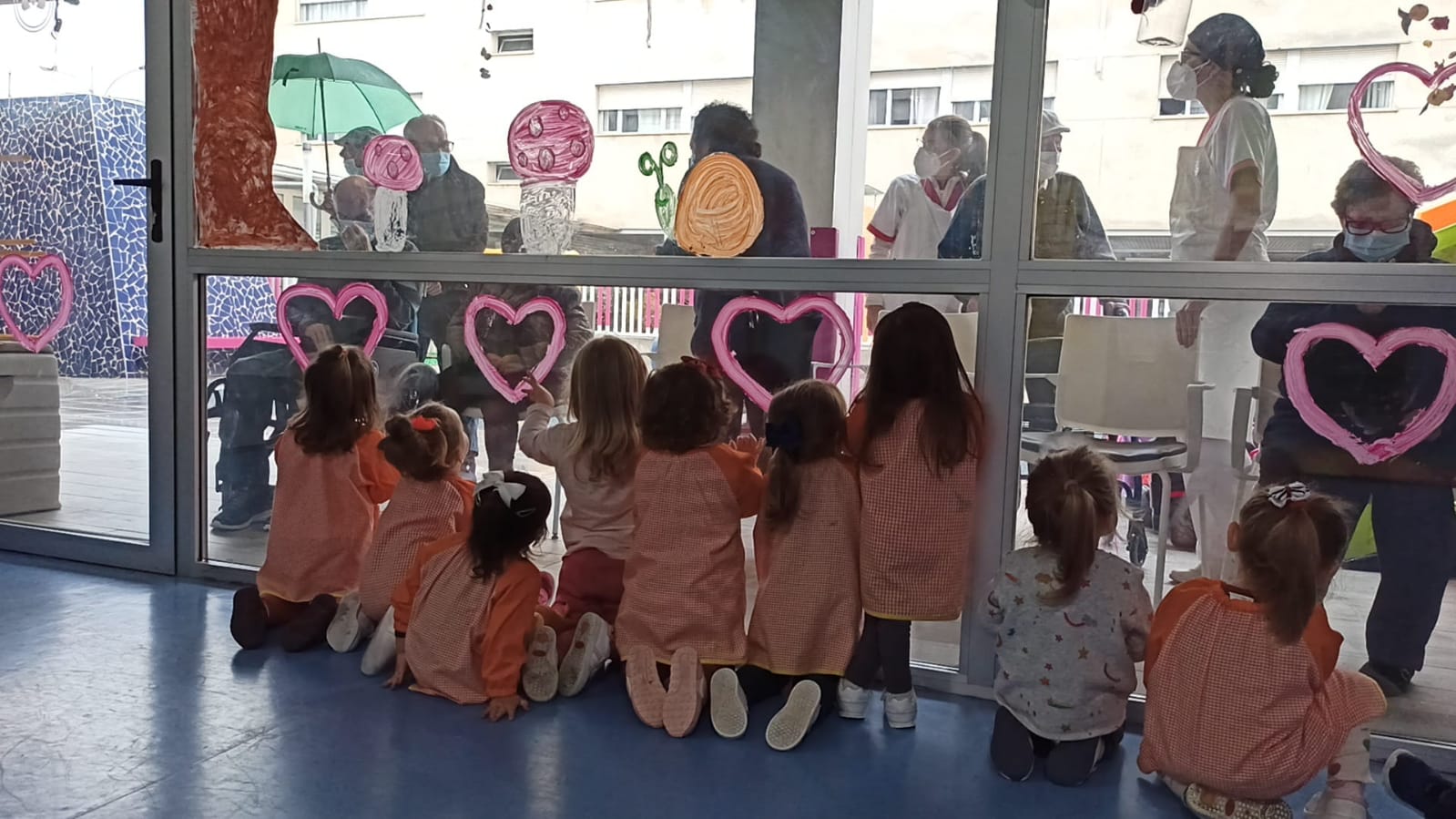 centro intergeneracional en el que un grupo de niños y niñas interactúan a través de un ventanal con un grupo de personas mayores